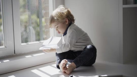 快乐的小男孩在家里的白色窗台上玩纸飞机