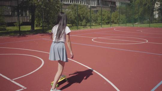 穿运动鞋裙子和T恤的女孩在户外篮球场上玩滑板