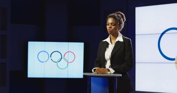 非裔美国主播在奥林匹克标志前阅读奥林匹克新闻，向左摇向一个制作团队