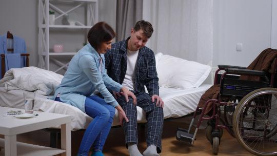 截瘫的高加索男子坐在床上，女人帮助穿上睡衣说话。幸福的丈夫和慈爱的妻子在睡前在家。家庭支持和护理概念。