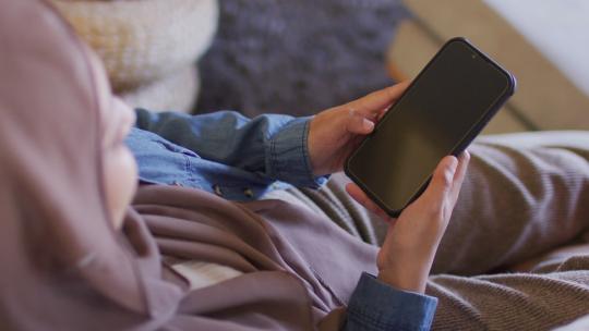 戴头巾的混血儿女人坐在家里使用智能手机的高角度视频，在屏幕上复制空间视频素材模板下载