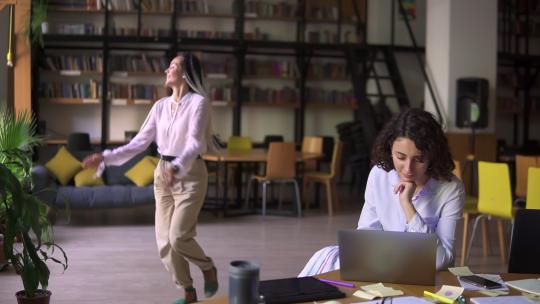 开放的公共图书馆。快乐的长发美丽的女孩穿着白色衬衫和米色裤子，在背景上滑稽地跳舞，而她的朋友则专注于学习，坐在桌旁用笔记本电脑工作