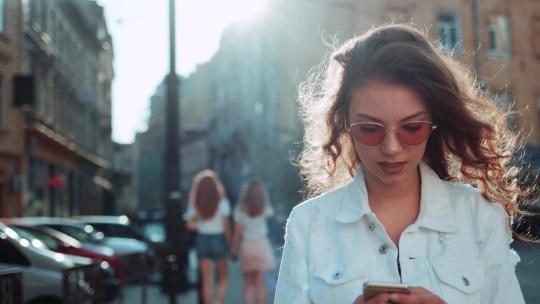 戴着时髦太阳镜的微笑卷发女人走在市中心的街道上，用着她的手机。穿着白色夹克的漂亮夏日女人走在街上，看着她的手机