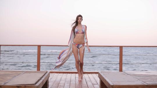 一个穿着比基尼和丝绸夏装的美丽模特走过木地板的慢动作镜头，背景是大海。风吹过，夏天