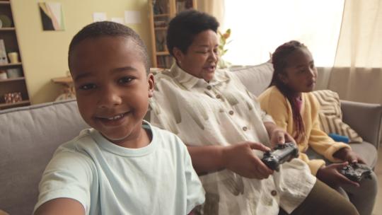 很酷的非裔美国青少年男孩录制自己在家里度过闲暇时间的视频，奶奶和姐姐坐在客厅的沙发上使用控制器玩电子游戏