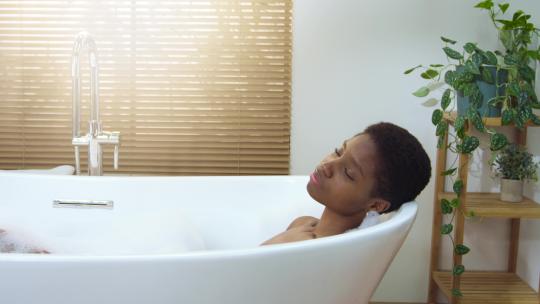 微笑的非洲迷人女人戴着耳机在浴室的泡沫浴缸里放松和跳舞