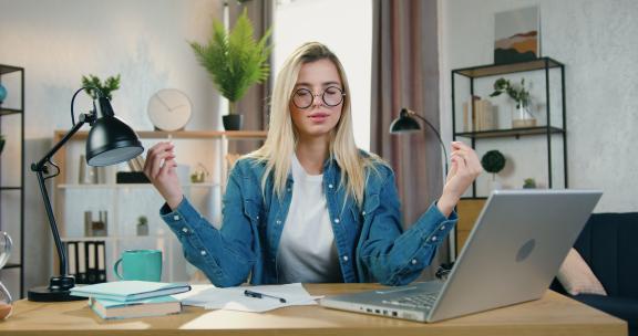 有吸引力的放松自信的年轻金发女子戴着眼镜，闭着眼睛冥想，打破她在家庭办公室笔记本电脑上的工作，工作流程和放松概念