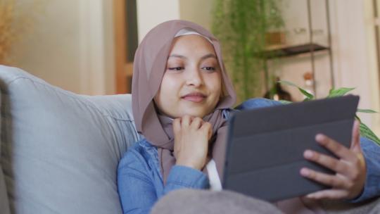 戴头巾的快乐混血儿女人在家里客厅的沙发上使用平板电脑放松的视频