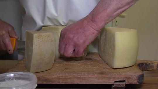 穿着白色围裙的奶酪制造商用巨型刀切开一块当地奶酪，向顾客提供，关闭